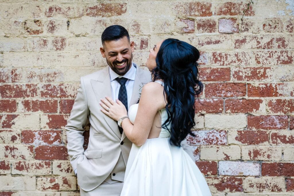 A smiling couple dressed in wedding attire stands against a weathered brick wall, with the woman touching the man's chest and both appearing happy. Drawn by Light Photography