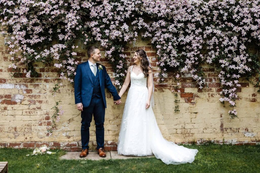 A bride and groom stand holding hands in front of a brick wall covered with blooming pink flowers, looking at each other and smiling. Drawn by Light Photography