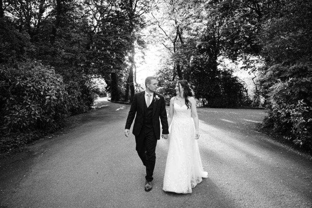 A bride and groom walk hand in hand down a tree-lined road, dressed in wedding attire, surrounded by sunlight and shadows. Drawn by Light Photography