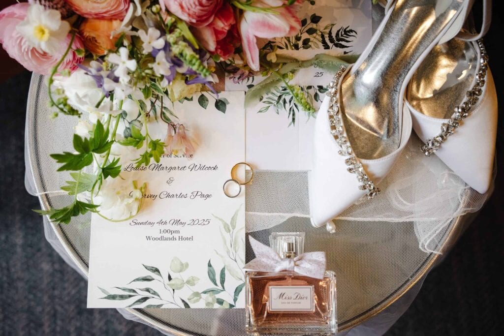 A flat lay of wedding items including an invitation, two gold rings, a pair of jewelled white shoes, a bouquet of flowers, and a bottle of Miss Dior perfume. Drawn by Light Photography