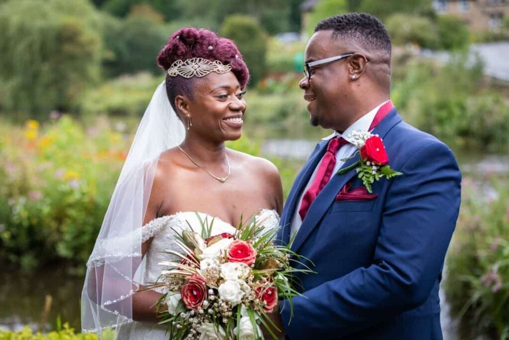 A bride and groom stand outdoors, smiling at each other. The bride holds a bouquet of red and white flowers, and the groom wears a navy suit with a red tie and buttonhole. Drawn by Light Photography