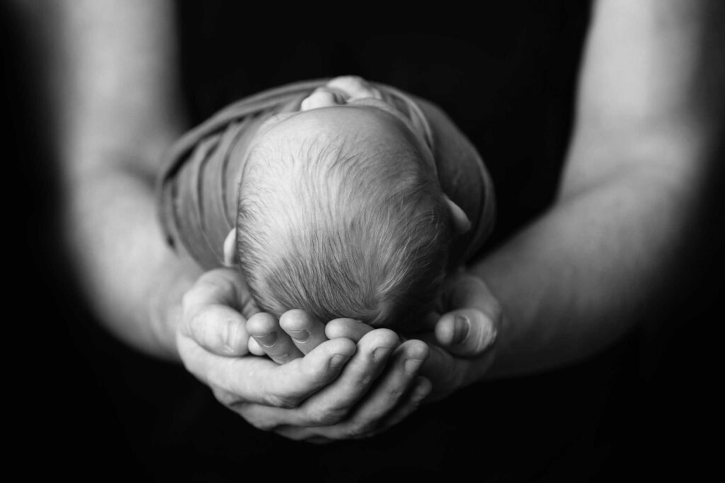 A close-up, black-and-white image of an adult holding a newborn baby gently in their hands against a dark background. Drawn by Light Photography
