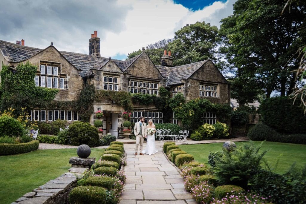 Two people in wedding attire walk down a stone path in a manicured garden in front of a large, historic, ivy-covered house. Drawn by Light Photography