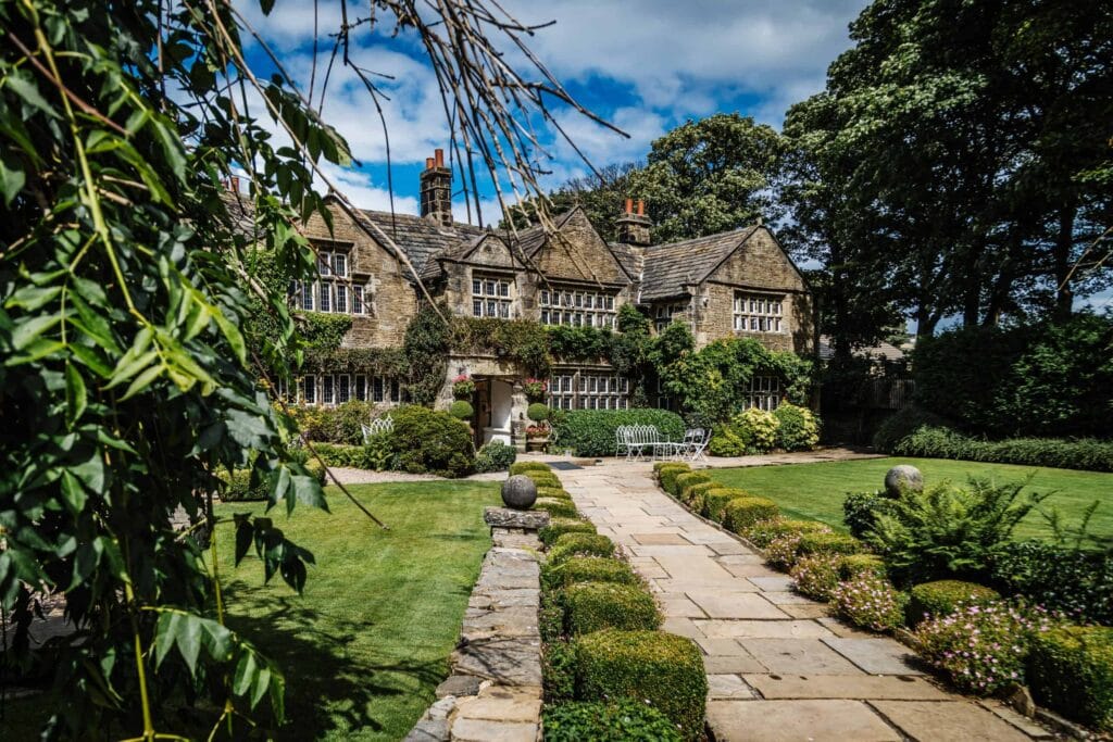 A stone path leads to a large, historic stone house with tall chimneys, surrounded by manicured gardens and greenery under a partly cloudy sky. Drawn by Light Photography