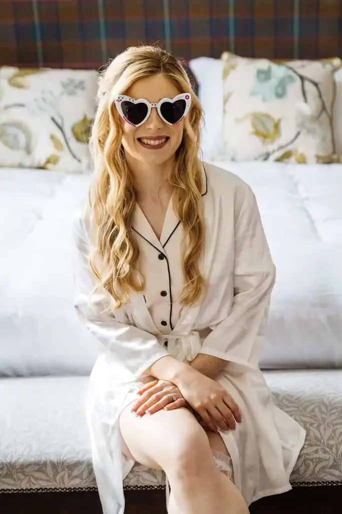 A woman with long blonde hair wearing heart-shaped sunglasses and a white dressing gown sits on the edge of a bed, smiling at the camera. Drawn by Light Photography