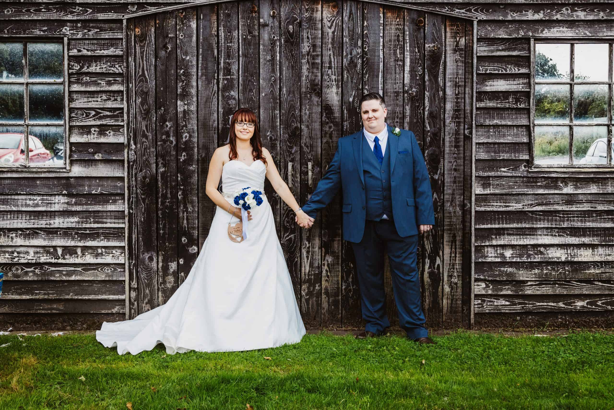 A bride in a white dress and a groom in a blue suit stand holding hands in front of a weathered wooden barn wall. Drawn by Light Photography