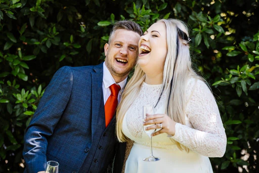 A man in a blue suit and red tie and a woman in a white dress are standing close together outdoors, both laughing and holding glasses of champagne. Drawn by Light Photography