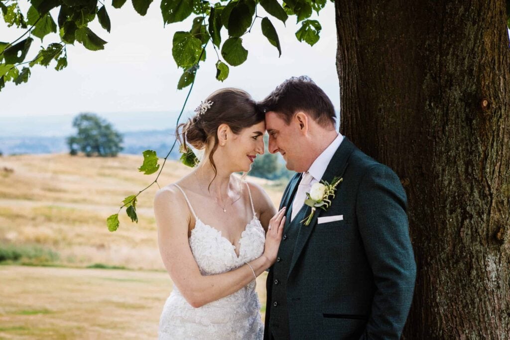 A bride and groom stand close together under a tree, touching foreheads and smiling, with a rural landscape in the background. Drawn by Light Photography