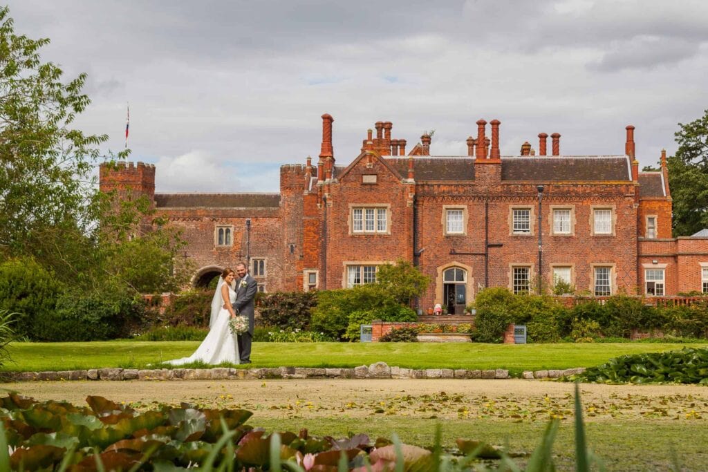 A bride and groom stand together in front of a large historic red-brick manor with gardens and a cloudy sky in the background. Drawn by Light Photography