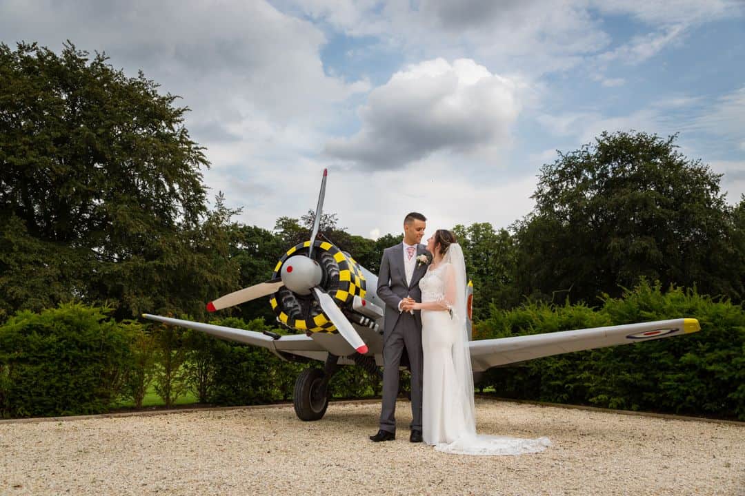 A bride and groom stand together in front of a vintage propeller aeroplane on a gravel path, with green bushes and trees in the background. Drawn by Light Photography