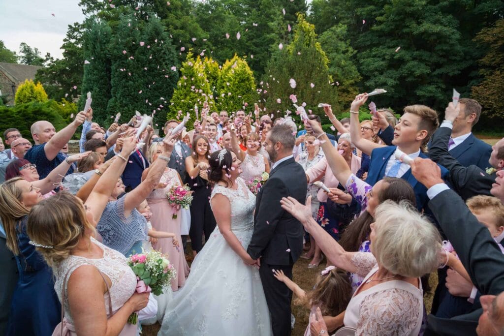 A bride and groom stand in the centre of a crowd outdoors as guests throw confetti and celebrate, surrounded by trees and greenery. Drawn by Light Photography