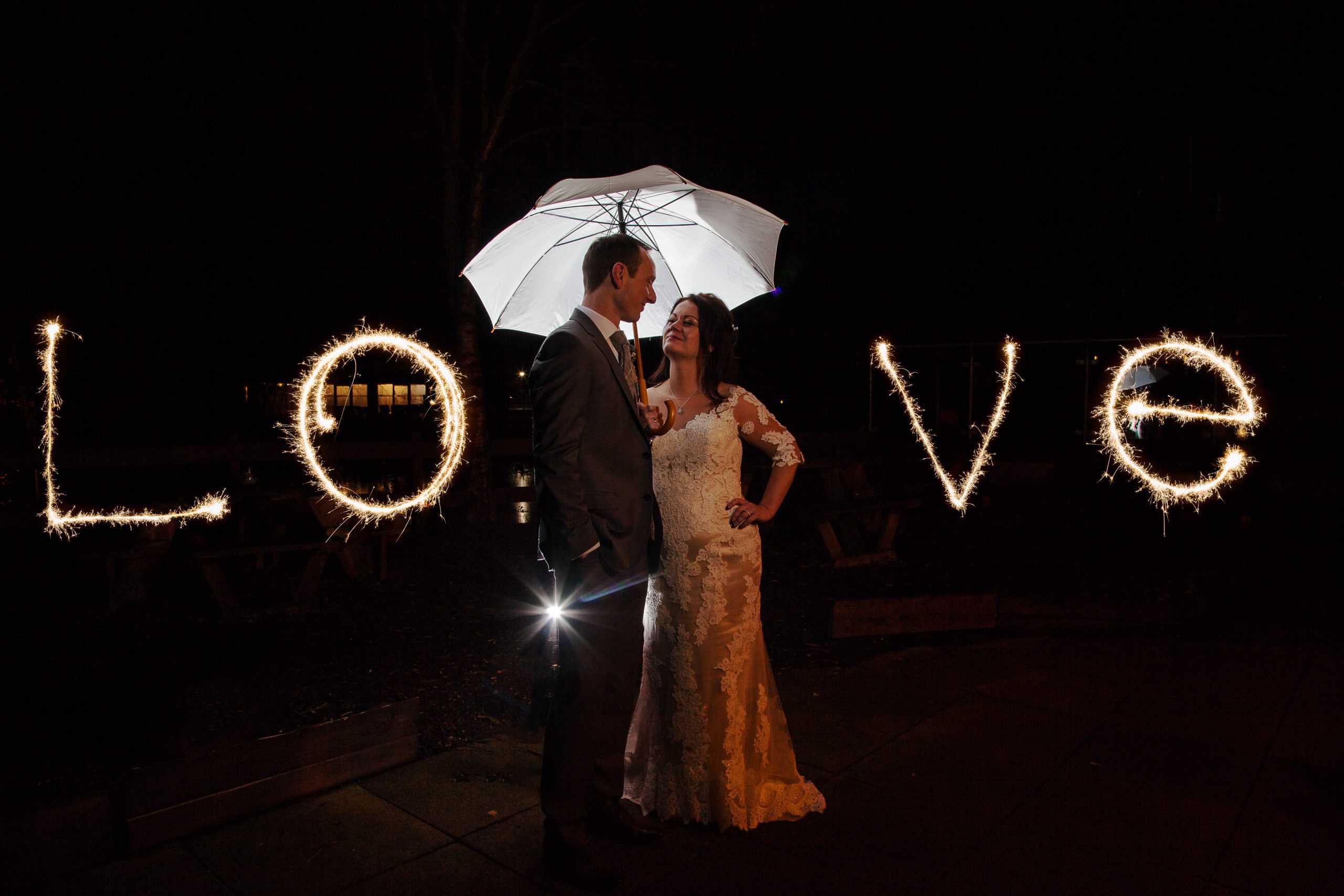 A bride and groom stand under a white umbrella at night, surrounded by the word "LOVE" written with sparklers. Drawn by Light Photography