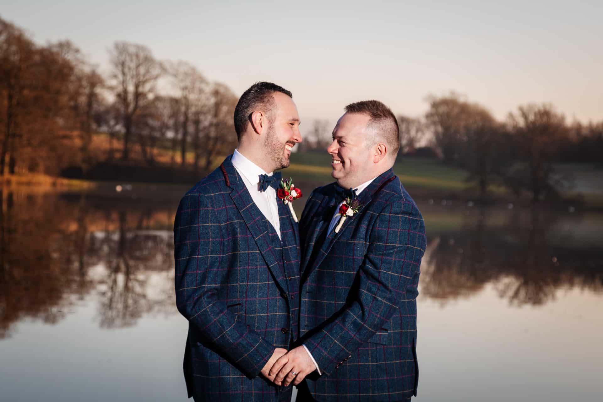 Two men in matching checked suits stand holding hands and smiling at each other by a calm lake with trees in the background. Drawn by Light Photography