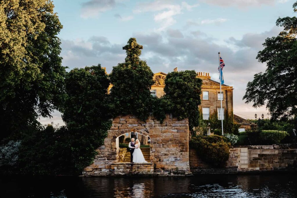 A bride and groom stand together in an ivy-covered stone archway by the water, with a large historic building and flags in the background. Drawn by Light Photography