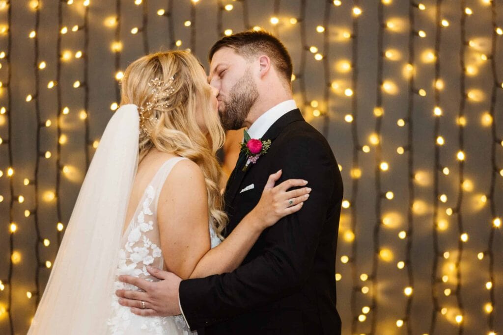A bride and groom share a kiss in front of a backdrop of fairy lights. Drawn by Light Photography