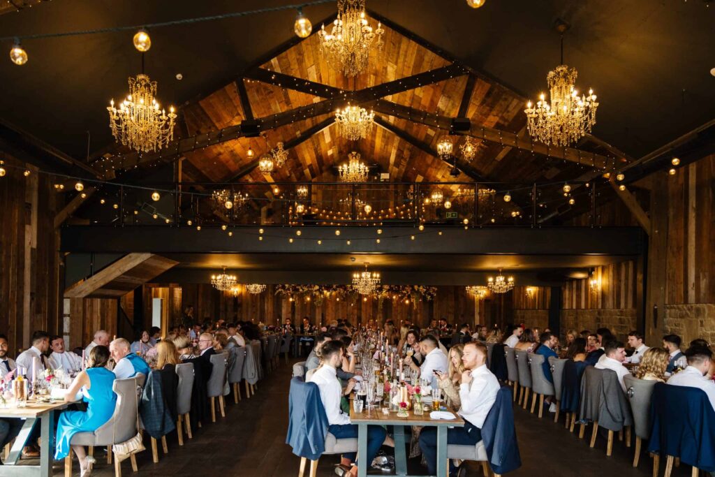 A large group of people dine at long tables in a rustic hall with chandeliers and wooden beams, under warm ambient lighting. Drawn by Light Photography