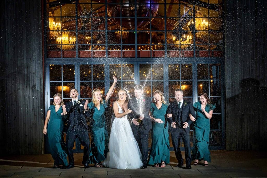 A wedding party poses in front of large windows at night, with members cheering as someone sprays champagne, creating an arc of droplets above them. Drawn by Light Photography