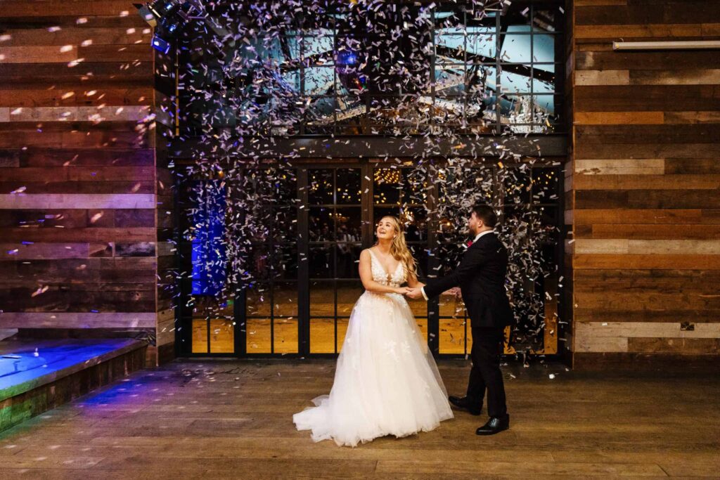 A bride and groom stand together indoors beneath falling confetti, holding hands and looking at each other, with wooden walls and glass doors in the background. Drawn by Light Photography