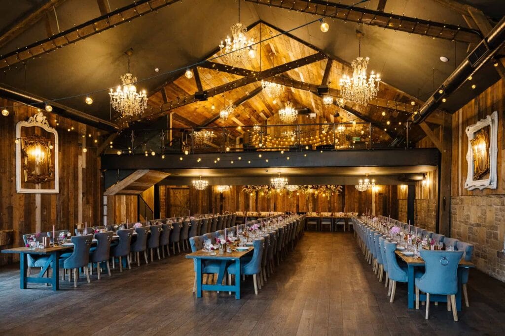 A rustic banqueting hall with wooden walls, chandeliers, fairy lights, and two long dining tables set for an event, with blue upholstered chairs on either side. Drawn by Light Photography