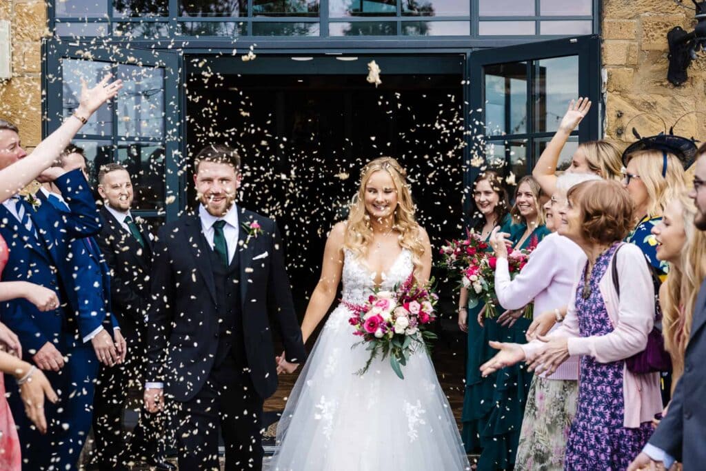 A bride and groom walk outside as guests throw confetti and celebrate; the bride holds a bouquet of flowers and both are smiling. Drawn by Light Photography