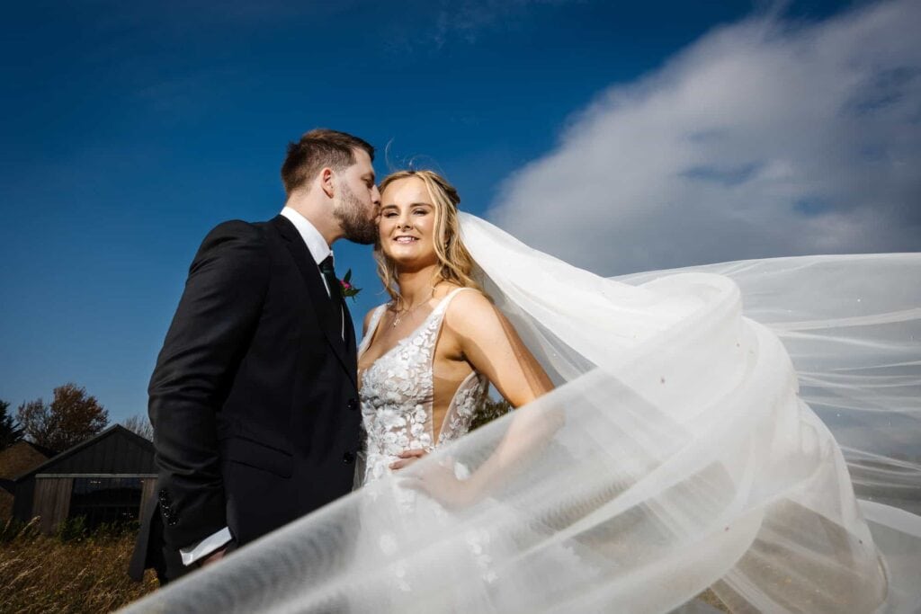 A groom in a black suit kisses the bride's temple as she smiles at the camera; her veil flows in the wind against a blue sky. Drawn by Light Photography