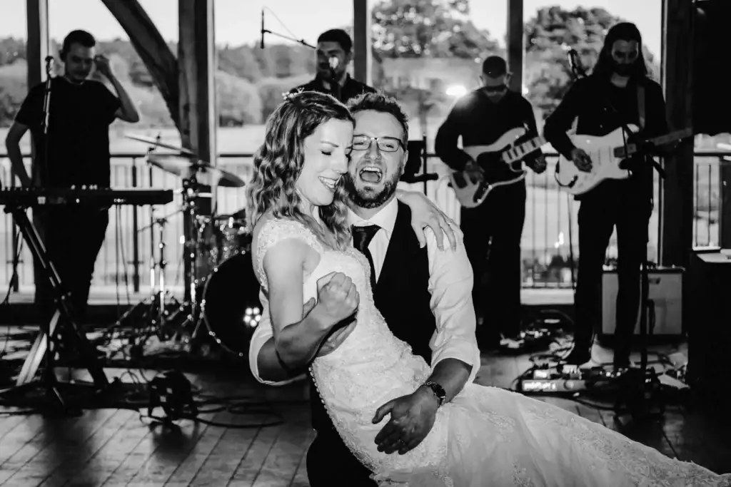 A groom lifts and smiles with a laughing bride on a dance floor, with a live band performing in the background. Drawn by Light Photography