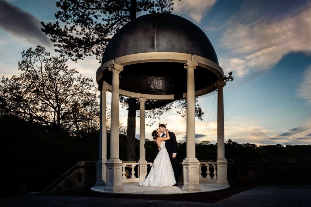 A bride and groom embrace under a domed pavilion at sunset with trees and a cloudy sky in the background. Drawn by Light Photography