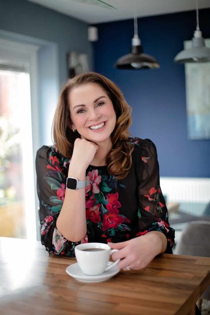 A woman with brown hair in a black floral blouse sits at a table, smiling, with her elbow on the table and a cup of coffee in front of her. Drawn by Light Photography
