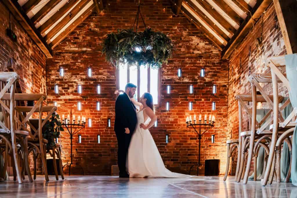 A bride and groom stand facing each other in a rustic brick venue with wooden beams, surrounded by candles and empty chairs. Drawn by Light Photography