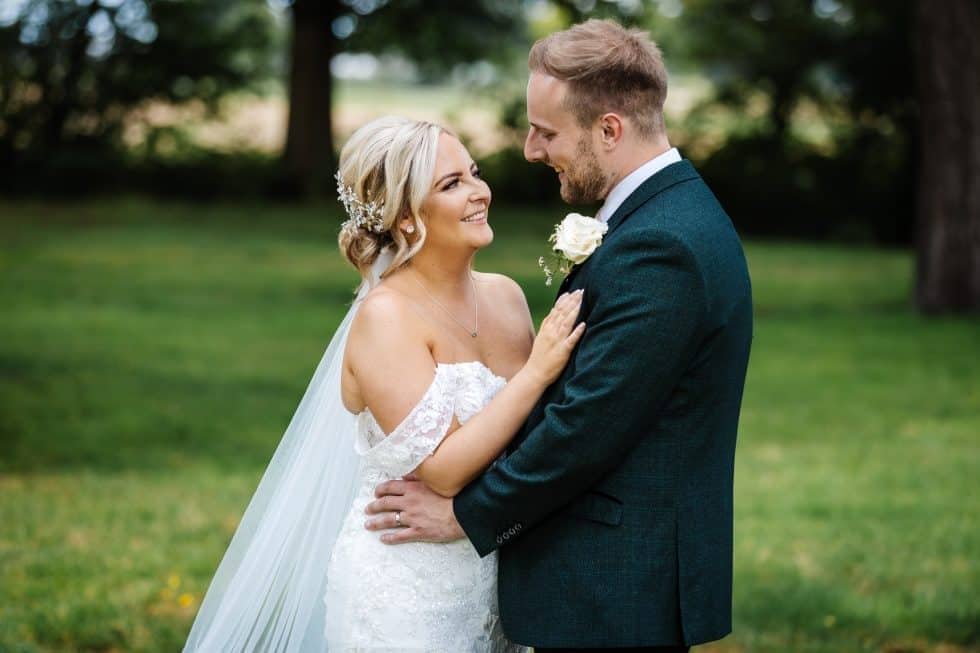 A bride and groom stand outdoors, facing each other and smiling, with greenery and trees in the background. The bride wears a white dress and veil; the groom wears a dark suit with a buttonhole. Drawn by Light Photography