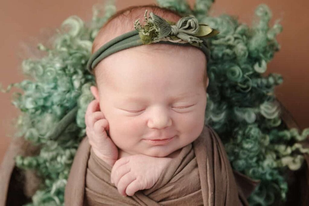 A newborn baby wrapped in brown cloth with a green headband, lying on curly green fabric, eyes closed and smiling softly. Drawn by Light Photography