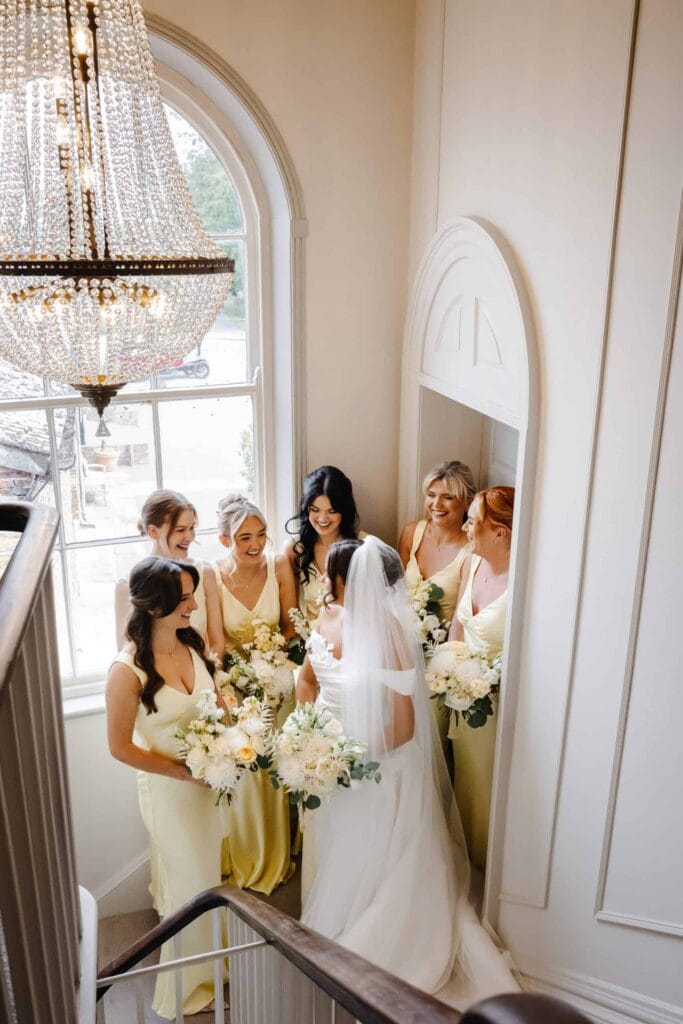 A bride in a white dress stands with five bridesmaids in light yellow dresses, all holding white bouquets, on a staircase beneath a large chandelier. Drawn by Light Photography