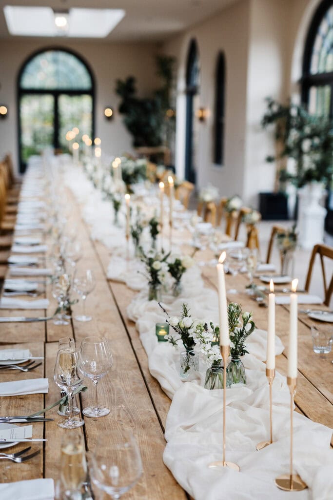 A long wooden table set for a formal event with white linen, candles, glassware, and floral centrepieces in a bright room with large arched windows, perfect for capturing by a Middleton Lodge Wedding Photographer. Drawn by Light Photography