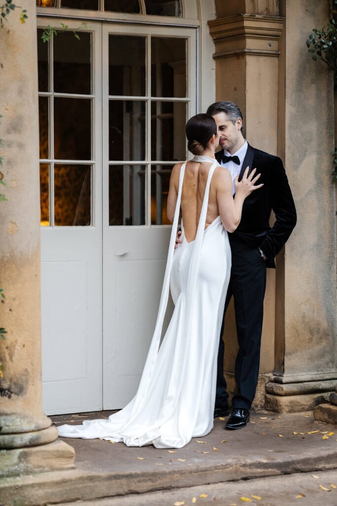 A woman in a white backless dress faces and touches a man in a black dinner jacket as they stand close together outside the elegant columns and glass doors of their Middleton Lodge wedding. Drawn by Light Photography
