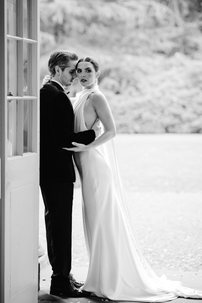 A man in a suit and a woman in a long, elegant gown stand closely together by an open door at a Middleton Lodge wedding, looking towards the camera. The image is captured in timeless black and white. Drawn by Light Photography