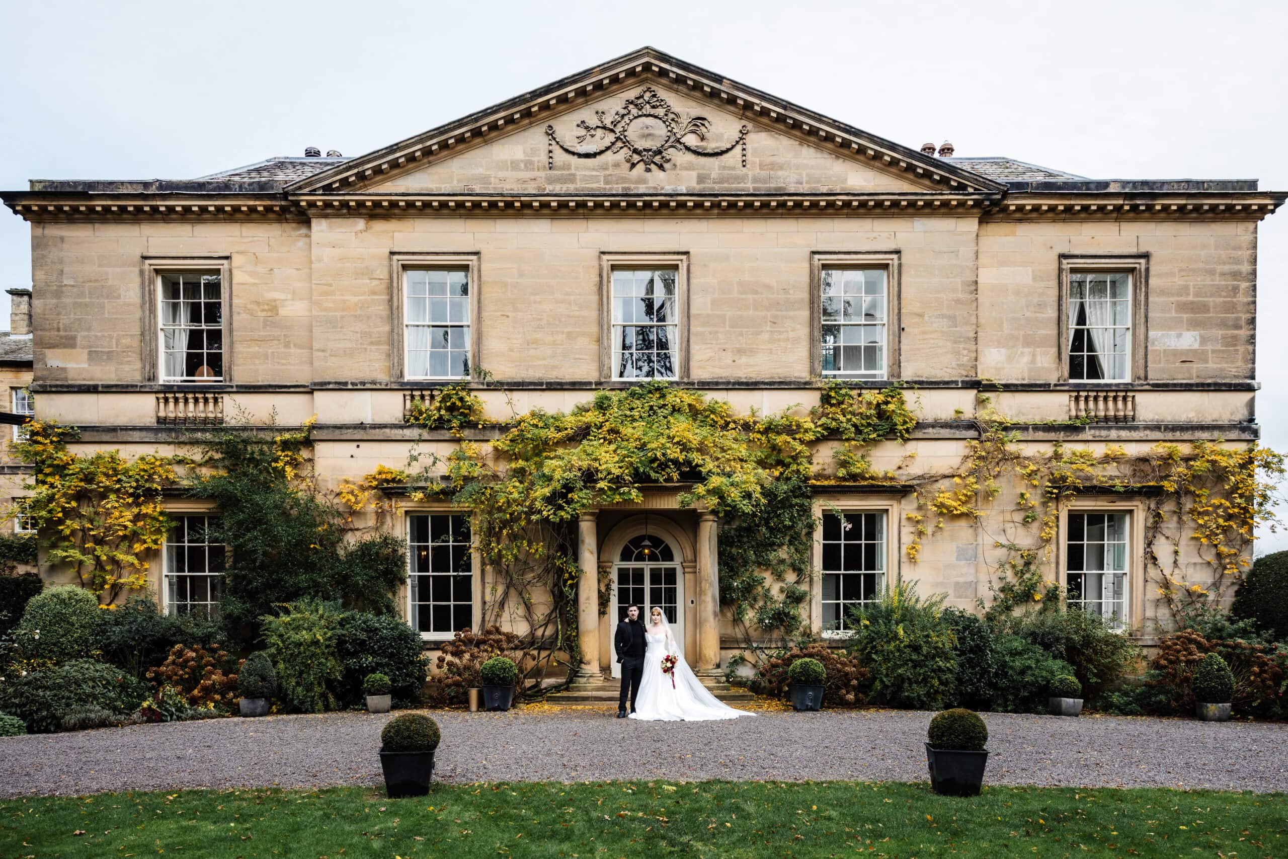 A bride and groom stand together in front of a large, historic stone manor house with ivy on the façade, beautifully captured by a Middleton Lodge wedding photographer. Drawn by Light Photography