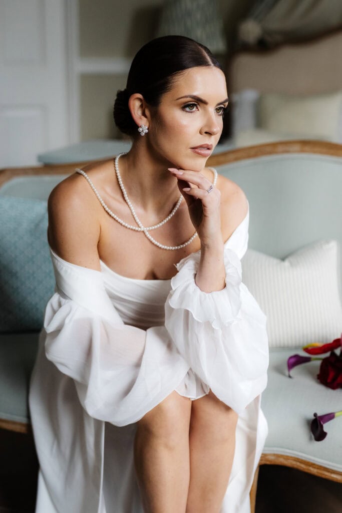 A woman in a white dress and pearl necklace sits on a light blue sofa, resting her chin on her hand and gazing thoughtfully ahead, evoking the timeless elegance of a Middleton Lodge wedding. Drawn by Light Photography