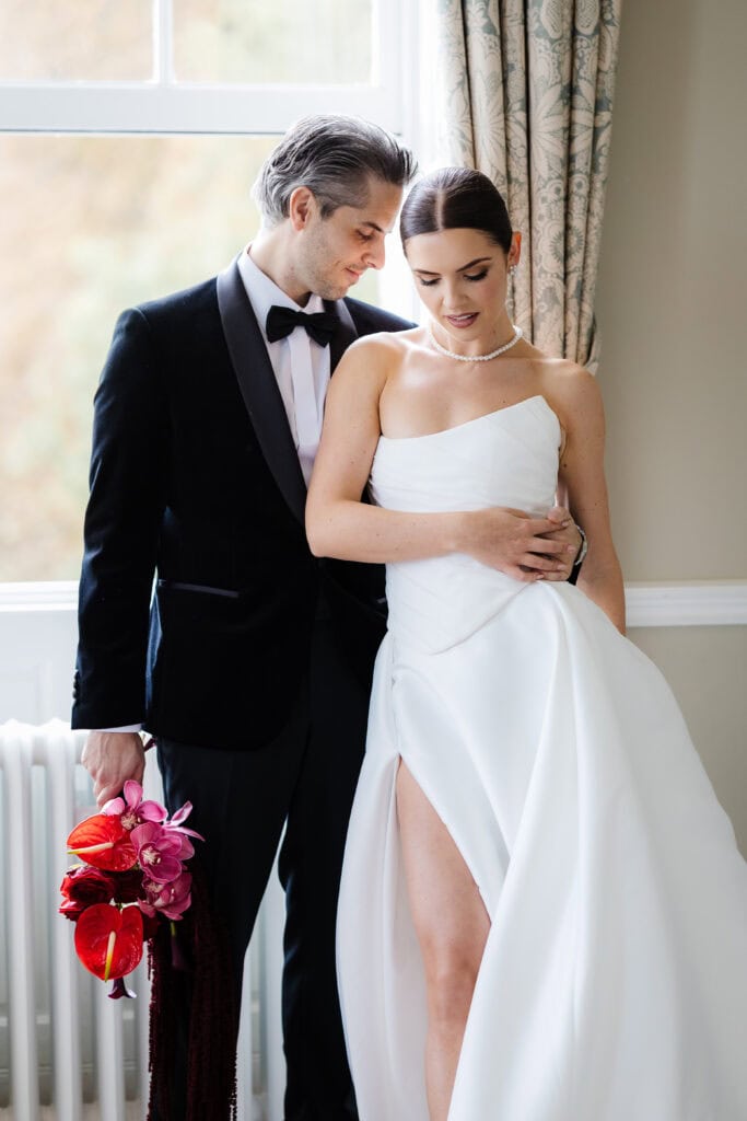 A man in a tuxedo stands beside a woman in a white wedding dress with a high slit, holding her waist. He holds a bouquet of pink and red flowers as they pose indoors by a window at their elegant Middleton Lodge wedding. Drawn by Light Photography