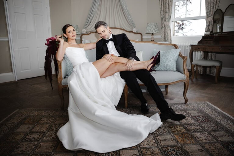 A bride in a white dress reclines with her legs across a groom in a black tuxedo as they sit on a vintage sofa in an elegant room at their Middleton Lodge wedding. Drawn by Light Photography