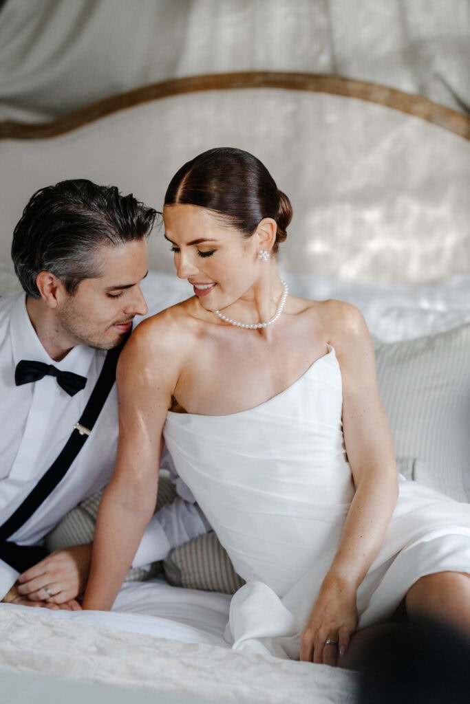A bride in a white dress and pearl necklace sits on a bed next to a groom in a white shirt and black bow tie, both appearing relaxed and content after their romantic Middleton Lodge wedding. Drawn by Light Photography