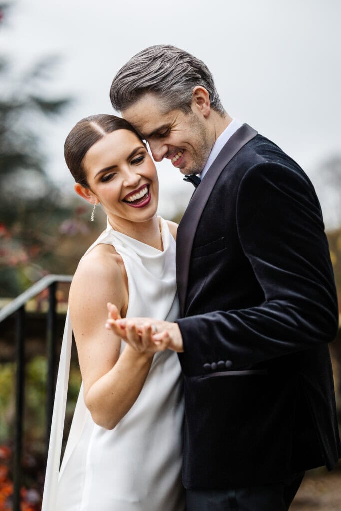A bride in a white dress and a groom in a dark suit smile and embrace outdoors at their Middleton Lodge wedding, holding hands. Drawn by Light Photography
