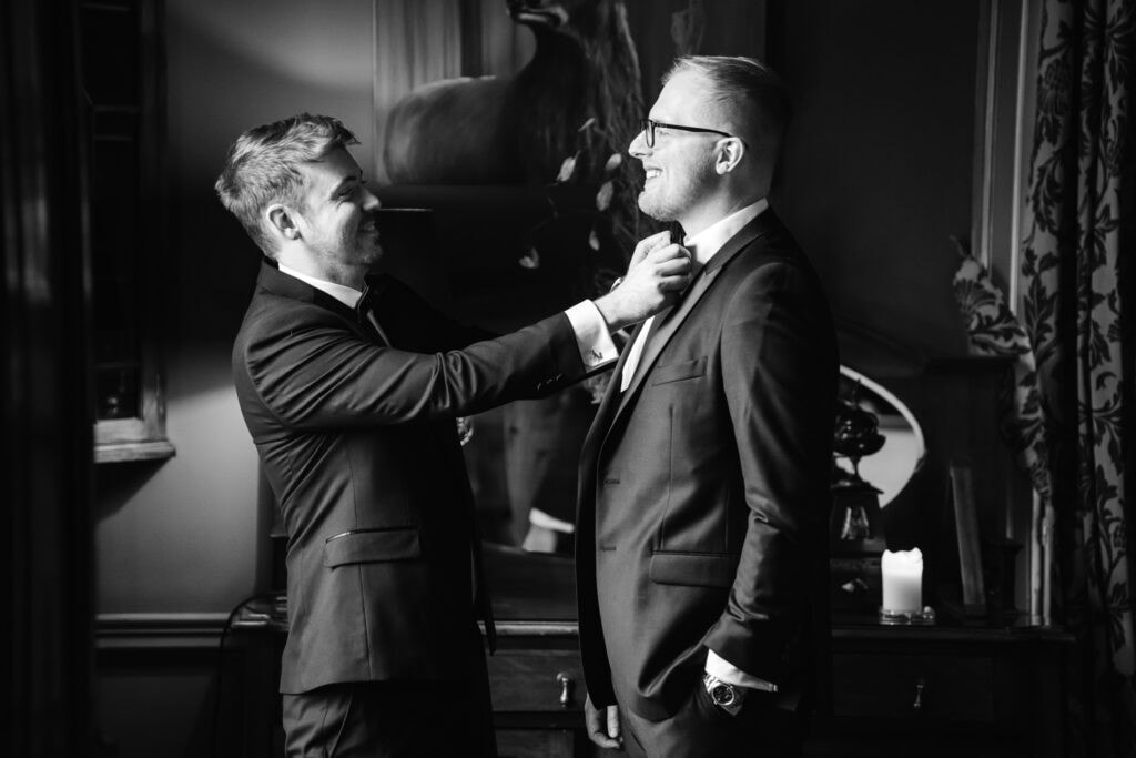Two men in suits stand indoors at a Middleton Lodge wedding; one adjusts the other's tie while both smile. A lit candle and decorative items are visible in the background. Drawn by Light Photography
