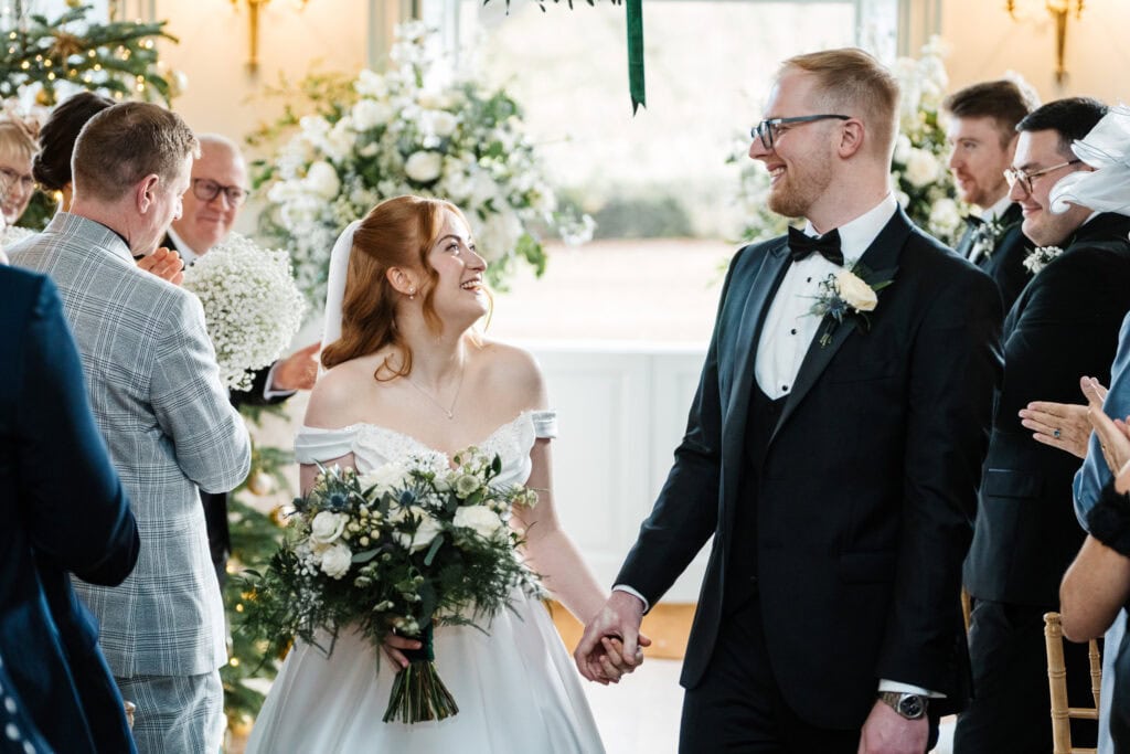 A bride and groom holding hands smile at each other whilst walking down the aisle at their Middleton Lodge wedding, surrounded by guests applauding in a beautifully decorated indoor setting. Drawn by Light Photography