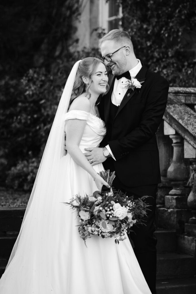 A bride and groom stand close together outdoors at their Middleton Lodge wedding, smiling. The bride holds a bouquet and wears an off-the-shoulder dress with a veil; the groom wears a tuxedo and glasses. Drawn by Light Photography