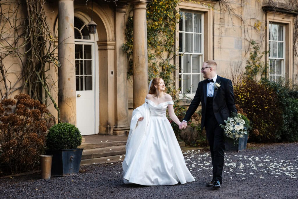 A bride in a white dress and a groom in a black suit hold hands and walk outside Middleton Lodge, a stone building beautifully decorated with greenery and flower petals. Drawn by Light Photography