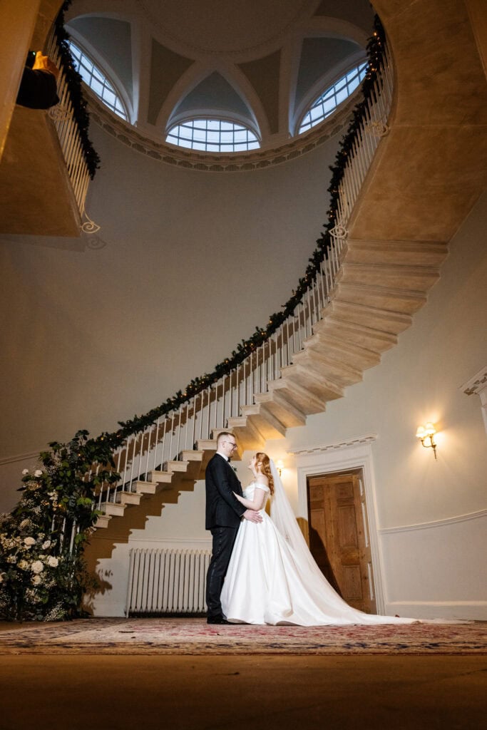 A bride and groom stand together beneath a curved staircase decorated with greenery in an elegant, high-ceilinged room at their Middleton Lodge wedding. Drawn by Light Photography