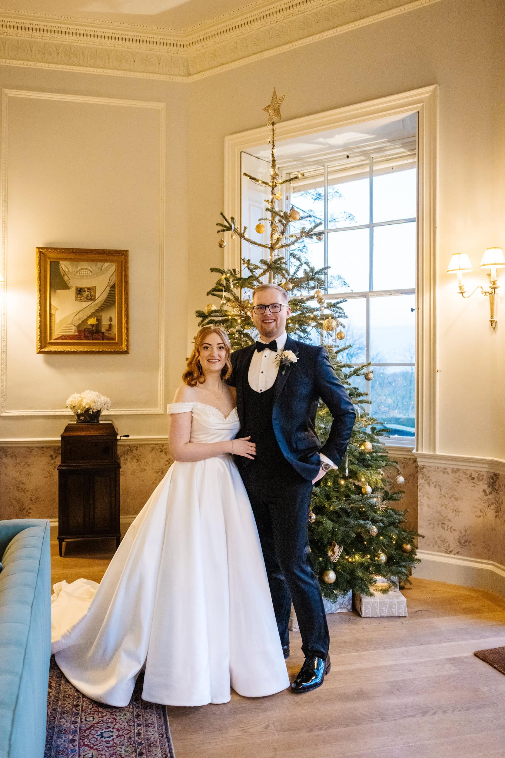 A bride and groom stand together in formal attire at their Middleton Lodge wedding, in a decorated room with a Christmas tree and large window in the background. Drawn by Light Photography