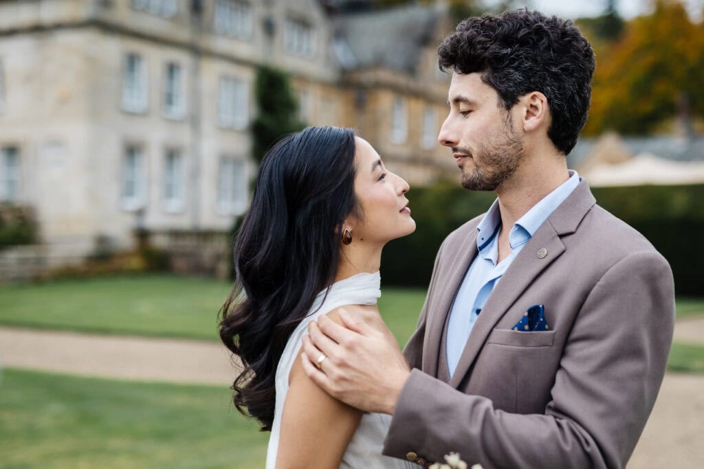 A couple stands close together outdoors, looking into each other's eyes, with a large building and greenery in the background. Drawn by Light Photography