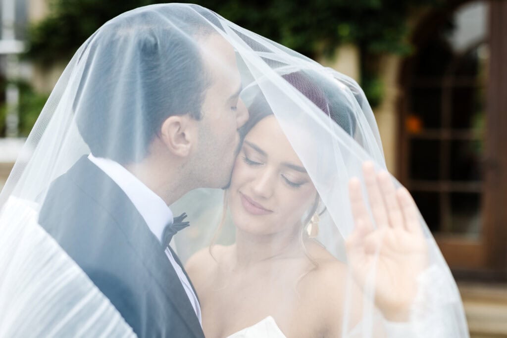 A groom kisses a bride on the forehead as they pose together under a translucent white veil, both dressed in formal wedding attire. Drawn by Light Photography