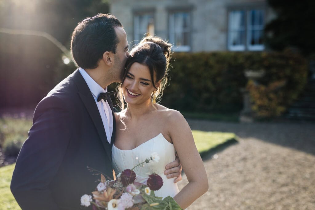 A groom in a suit kisses the bride on the forehead as they stand outside, smiling and holding a bunch of flowers. Drawn by Light Photography